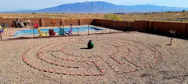 A backyard with a gravel area featuring a brick labyrinth pattern, a green vase at its center, and a swimming pool with…