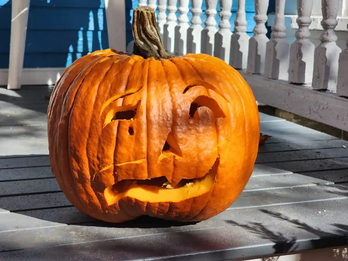 A carved orange pumpkin with a mischievous, asymmetrical face sits on a gray outdoor table, with a white picket fence and…