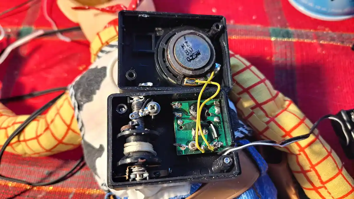 A close-up photo shows the open internals of a small electronic device, revealing a speaker, circuit board, and wires…