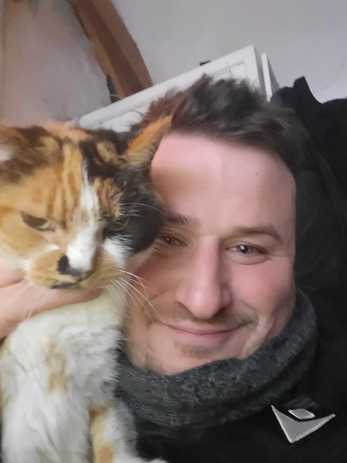 A person with dark hair and a grey scarf smiles at the camera while holding a calico cat. The cat, with orange, black, and…