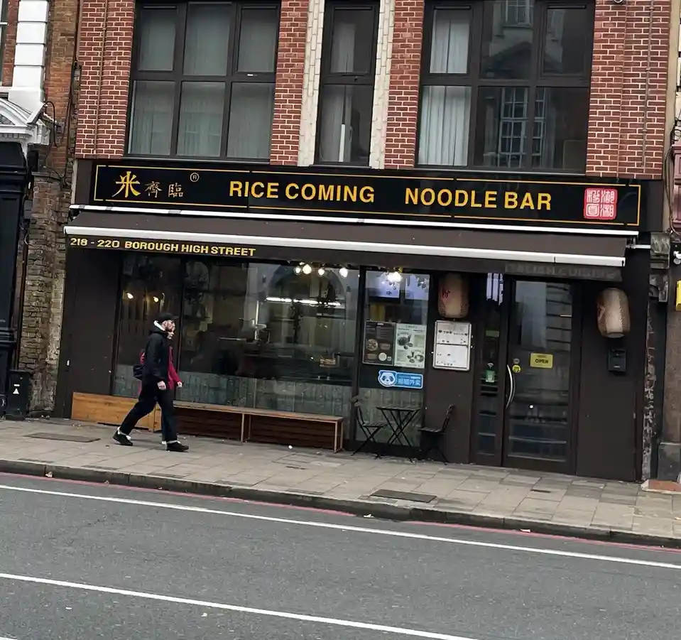A street-level photo shows a restaurant with a black sign that reads 'RICE COMING NOODLE BAR' in yellow letters, alongside…