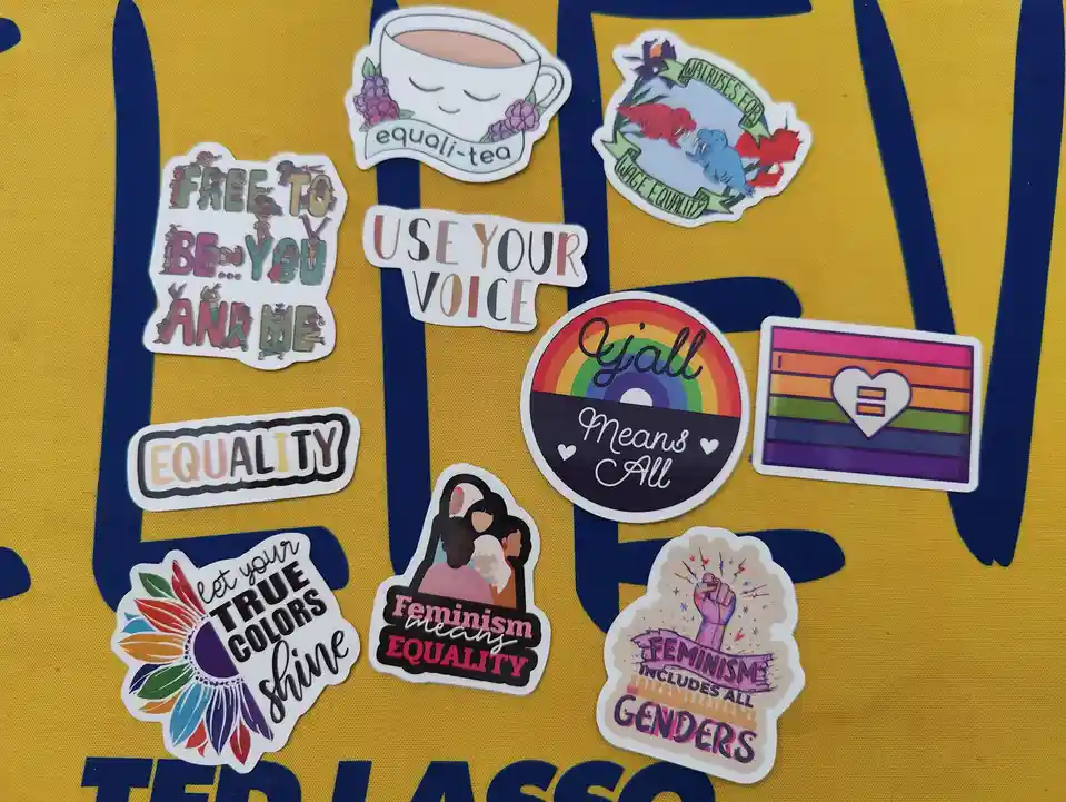 A collection of colorful feminist and equality-themed stickers arranged on a yellow fabric with blue lettering, including…