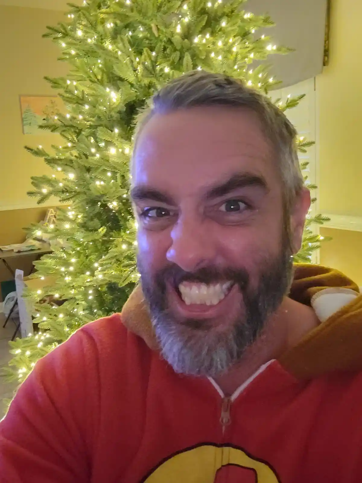 A man with a gray beard and mustache, wearing a red zip-up hoodie with a yellow emblem, smiles broadly at the camera while…