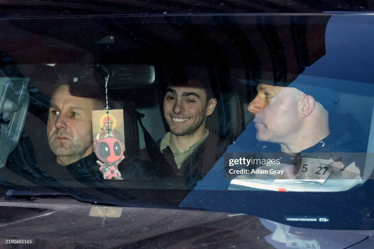 Three men are visible inside a car at night, viewed through the windshield. The driver and front passenger are looking…