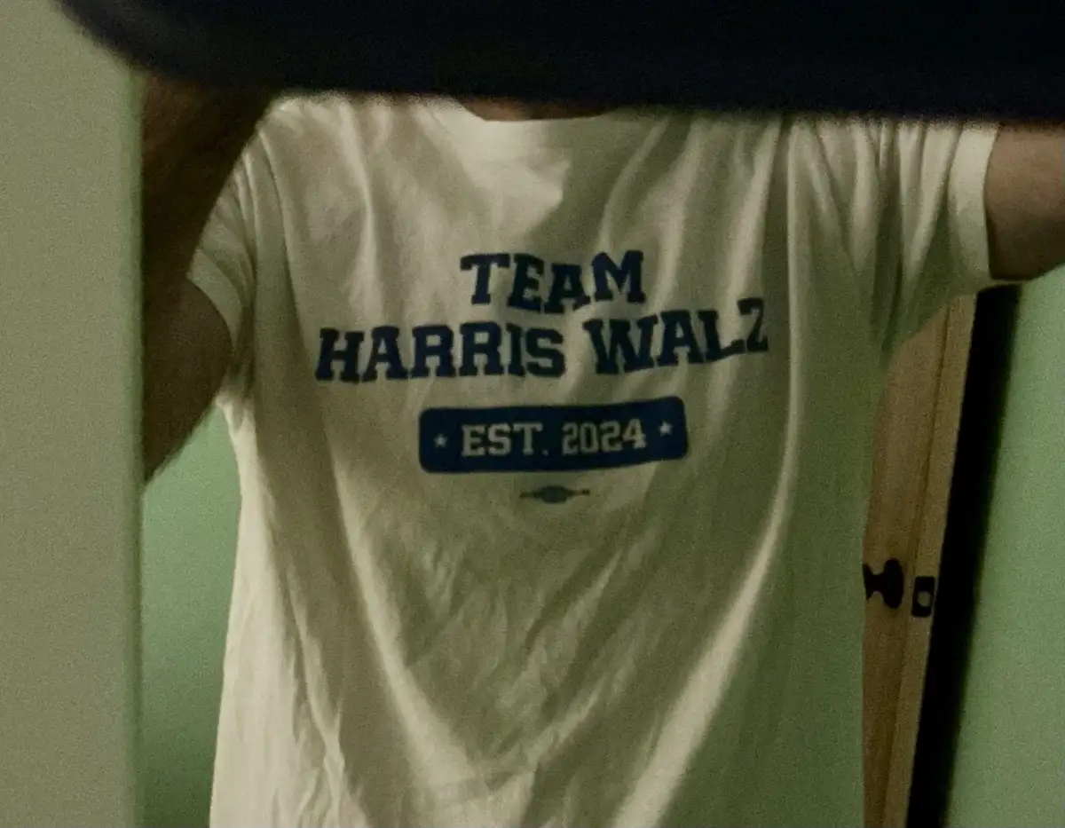 A person wearing a white t-shirt with blue text that reads 'TEAM HARRIS WALZ' and 'EST. 2024' in a blue rectangular box…