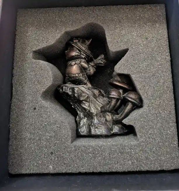 A small, dark bronze figurine of a fantasy character wearing a hat and holding a weapon, sitting atop a rocky base with…