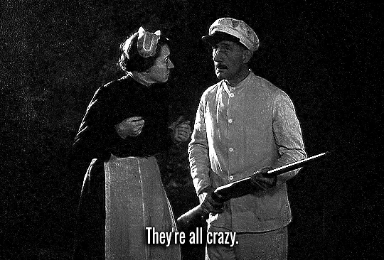 A black-and-white image shows a man holding a rifle and a woman standing beside him, both appearing to be in a tense…