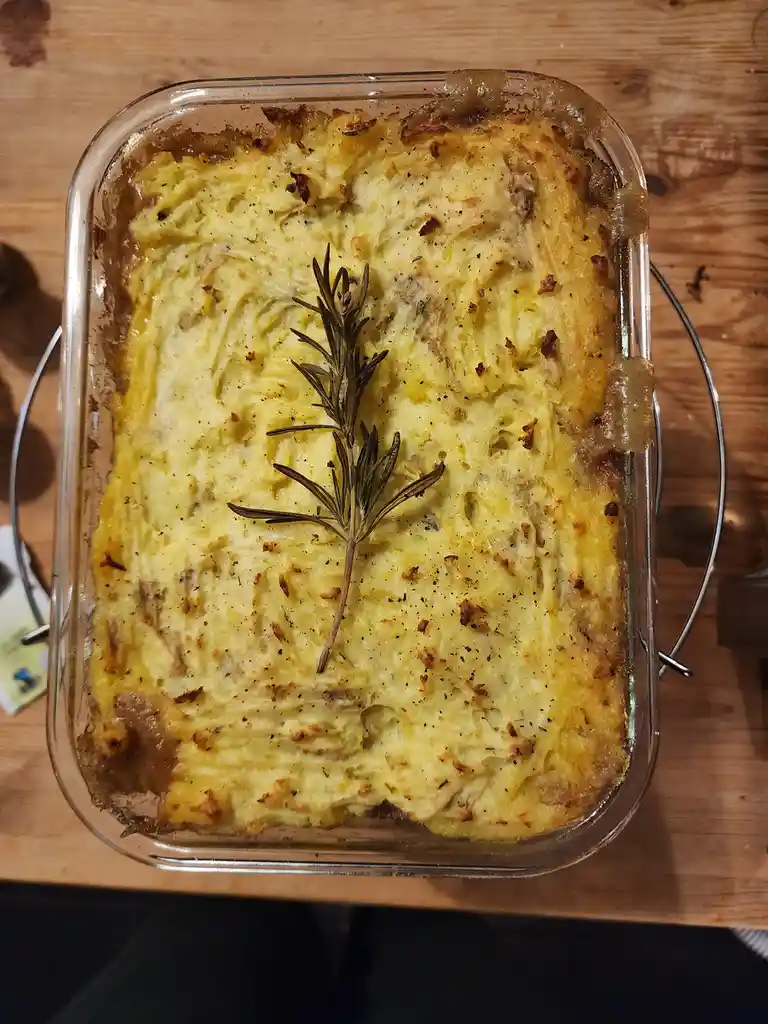 A top-down photo of a freshly baked shepherd's pie in a clear glass baking dish, resting on a wooden surface. The pie has a…