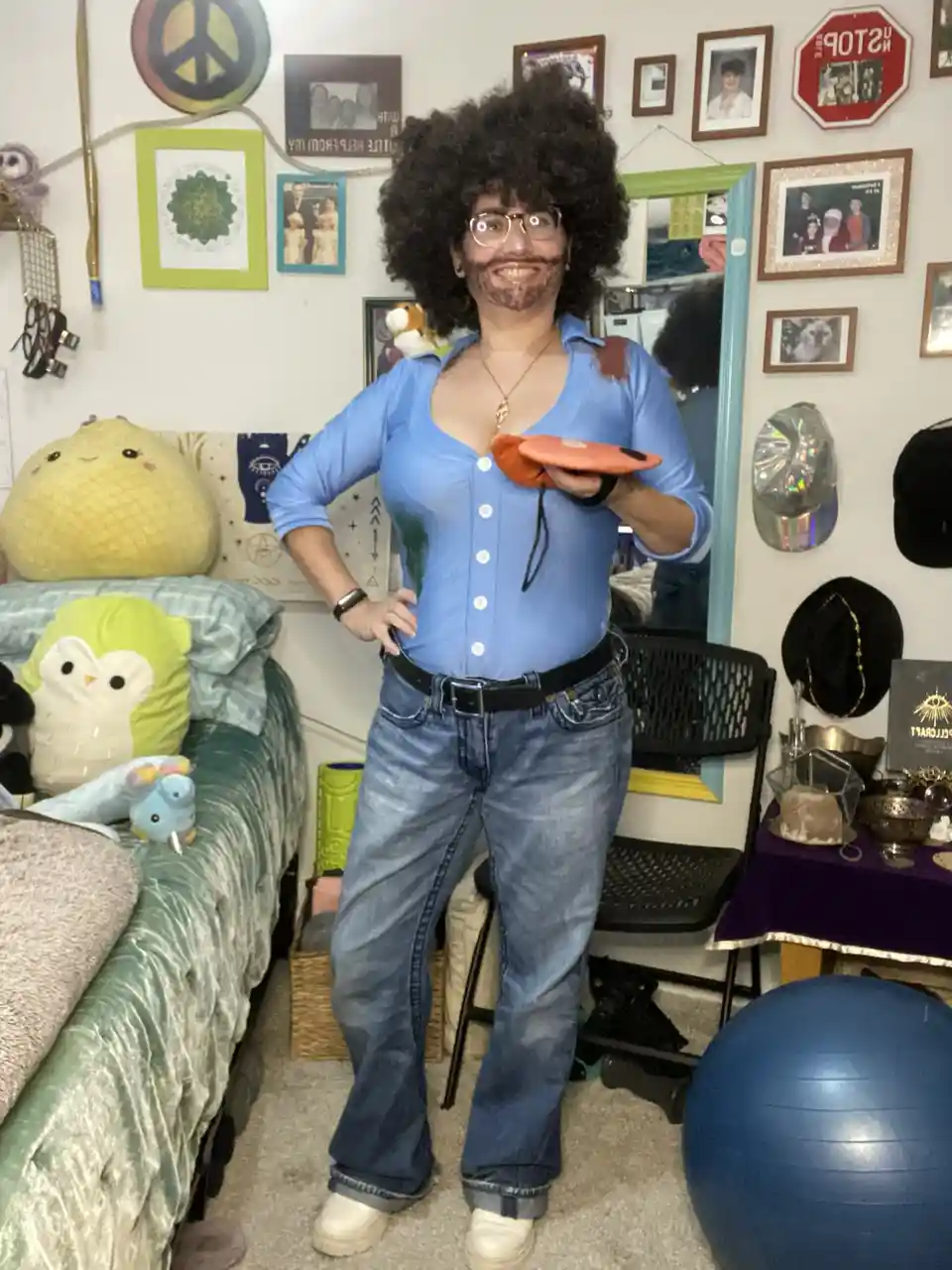 A person with a large afro wig, glasses, and a fake mustache stands in a bedroom holding a frisbee, wearing a blue button-up…