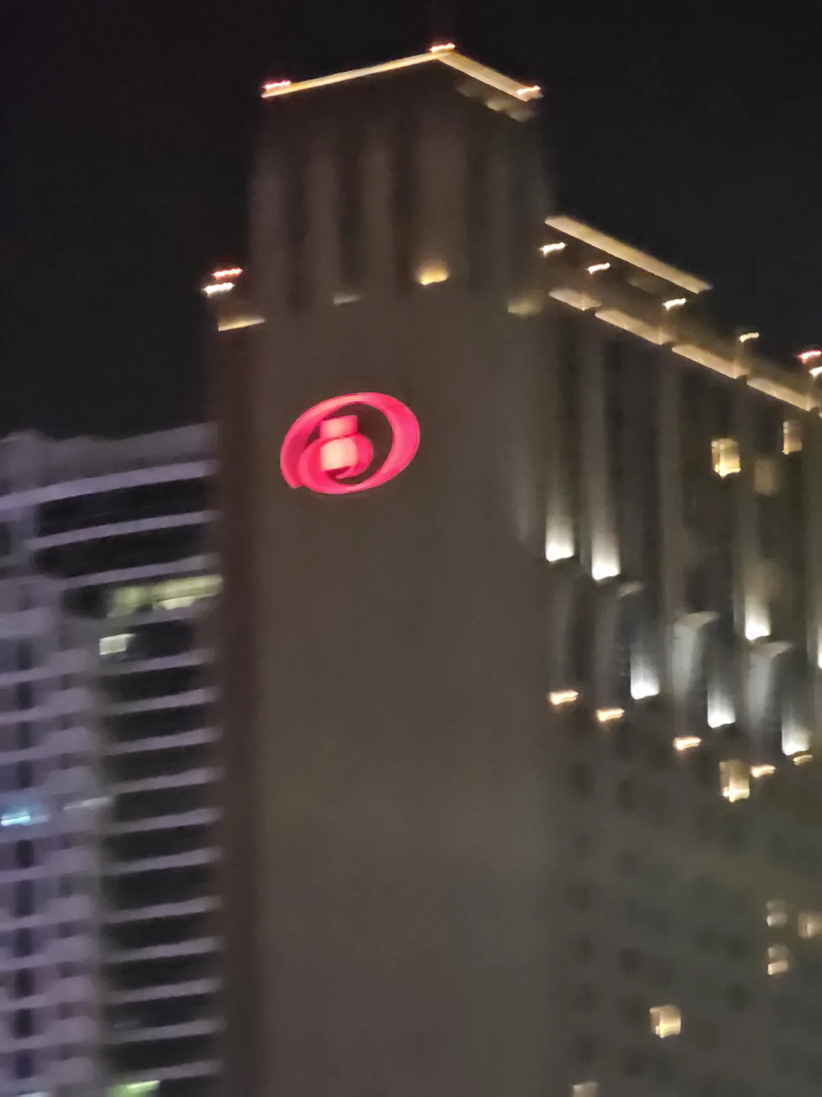 A blurry nighttime photo of a tall building with a glowing red circular logo on its facade, which resembles the Hilton hotel…
