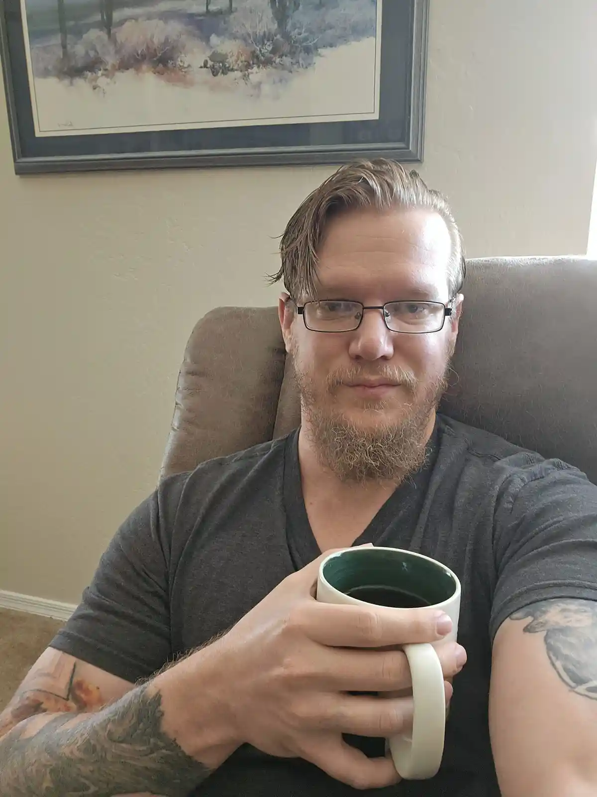 A man with glasses and a beard sits in a chair holding a mug, looking directly at the camera with a neutral expression. He…