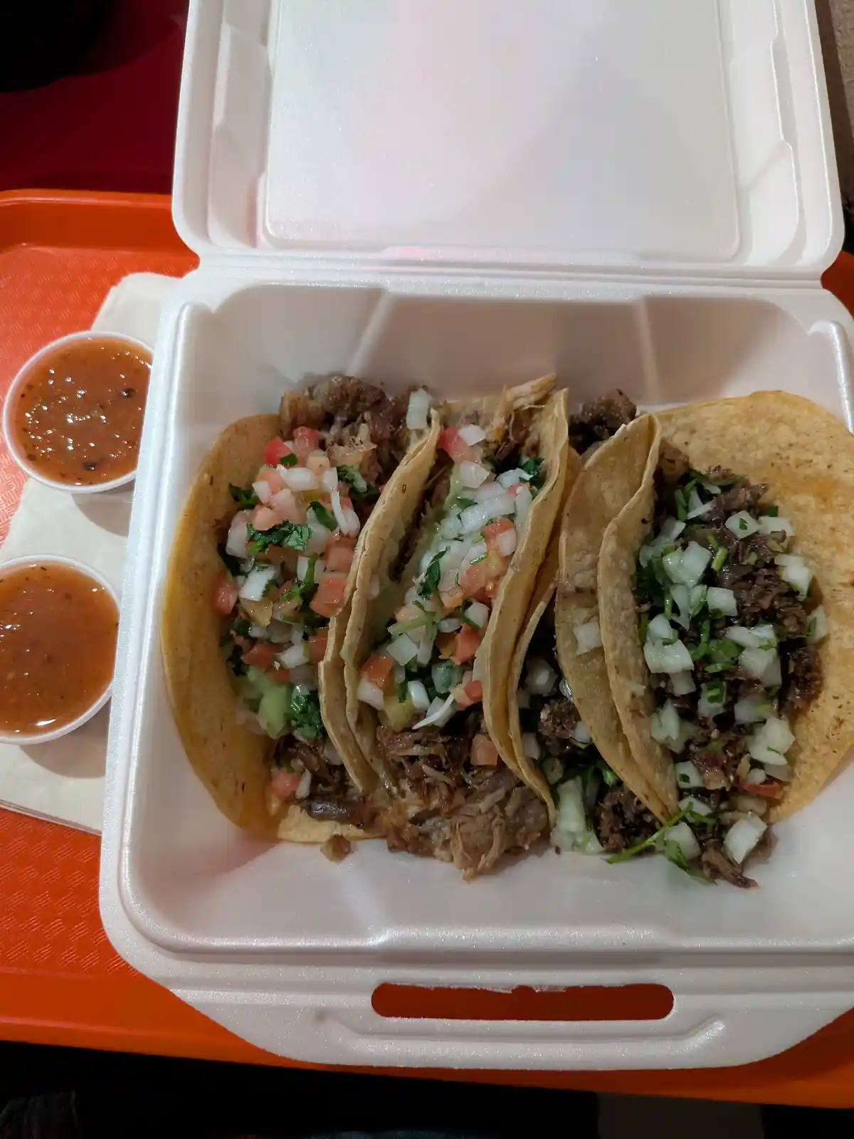 A white styrofoam takeout container holds three soft tacos filled with shredded meat and chopped onions, tomatoes, and…