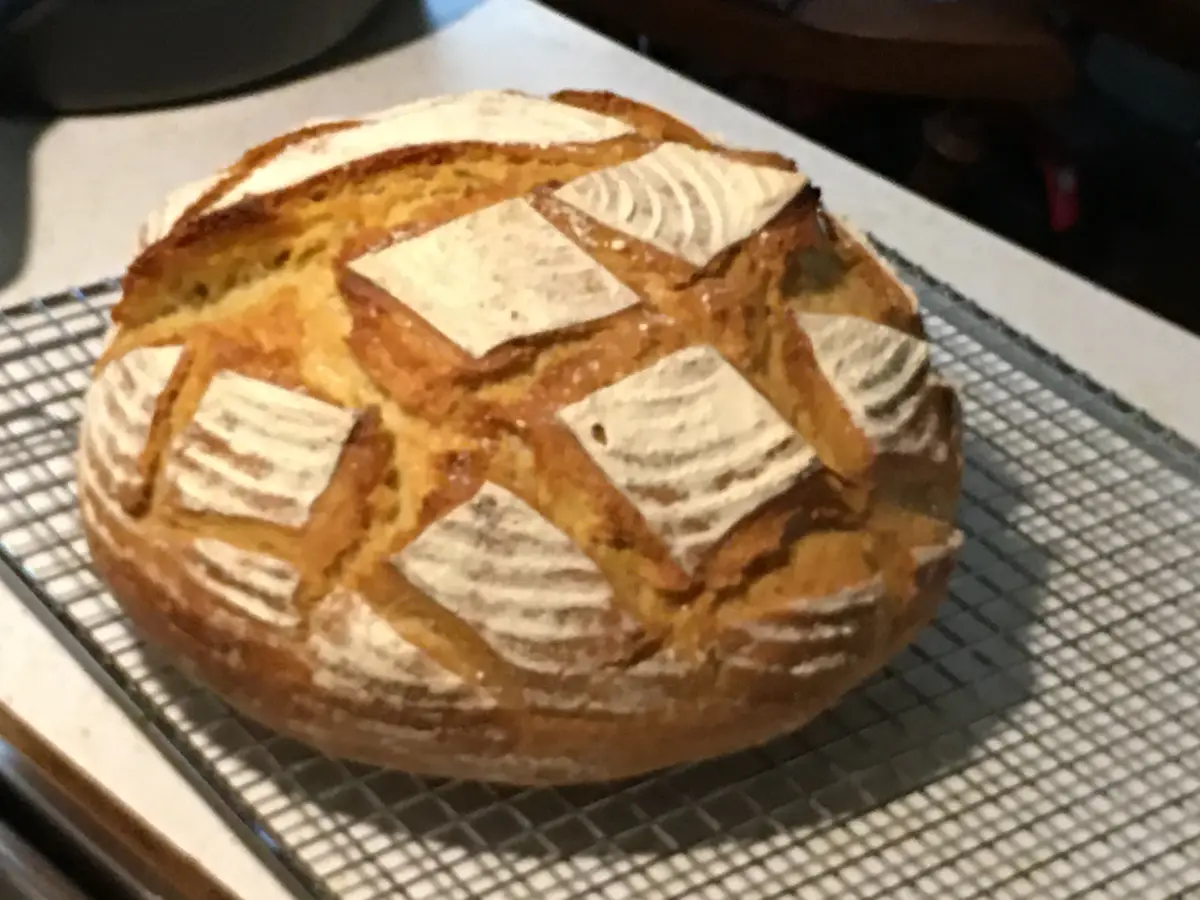 A round, golden-brown artisan bread loaf with a scored pattern sits on a metal cooling rack. The bread has a crusty exterior…