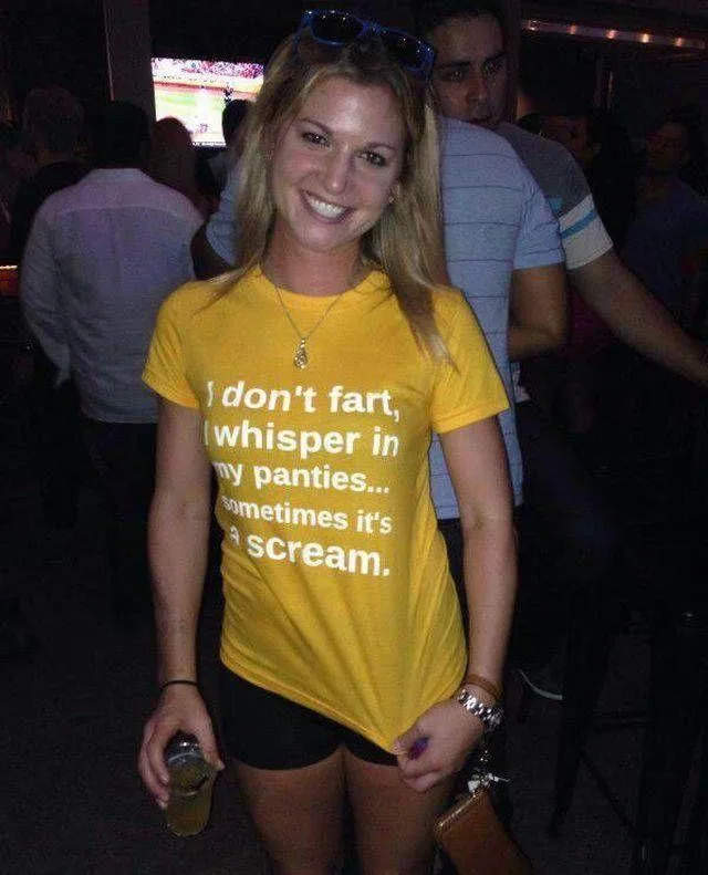A smiling blonde woman in a yellow t-shirt with white text stands in a dimly lit bar or club, holding a drink. The text on…