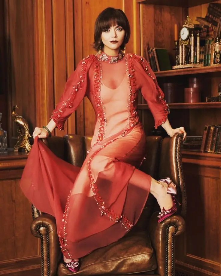 A woman with short dark hair sits in a brown leather armchair in a wood-paneled room with bookshelves. She wears a sheer…