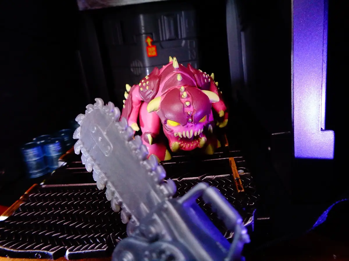 A pink, spiky, monstrous action figure with yellow eyes and teeth is positioned behind a chainsaw in the foreground, set…