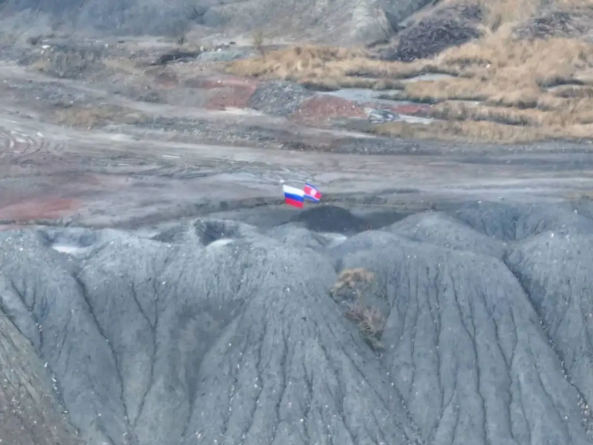 An aerial view of a barren, rocky, and excavated landscape with two small flags planted side-by-side in the middle ground…