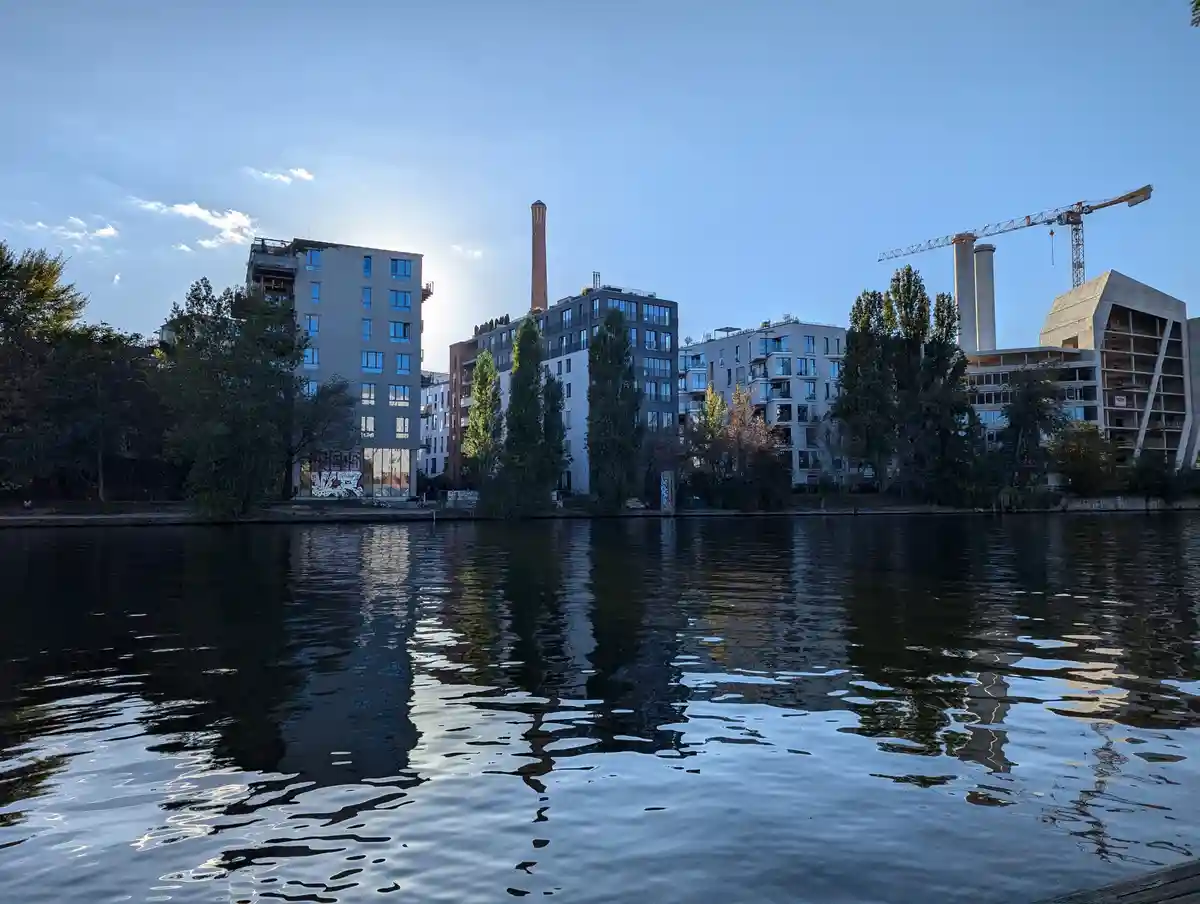 A scenic view of modern apartment buildings and industrial structures along a calm river, with a crane visible and the sun…