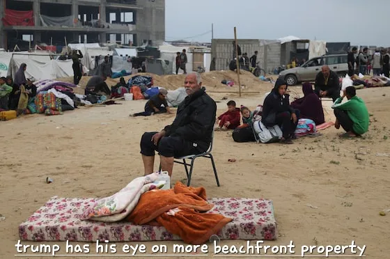A man sits on a folding chair in a sandy, makeshift camp with tents and people in the background. In front of him is a…