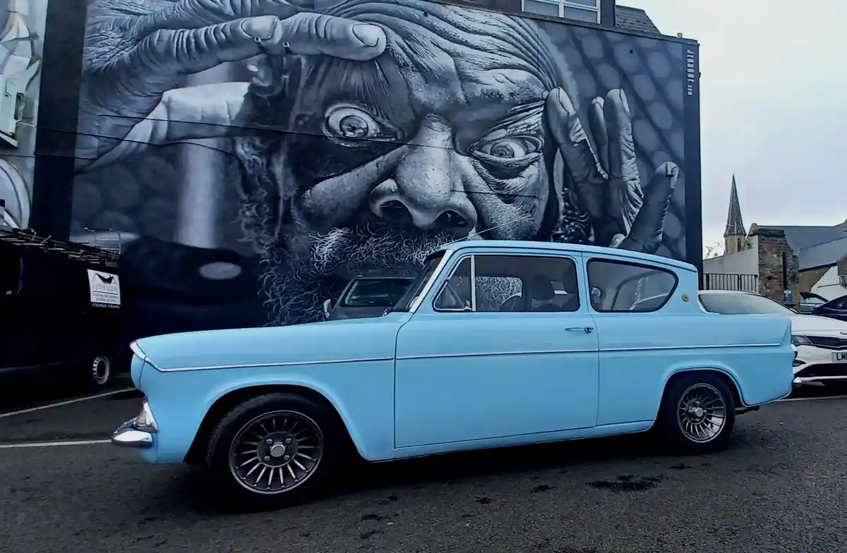 A light blue vintage car is parked in front of a large black-and-white mural of an elderly man with a shocked expression…