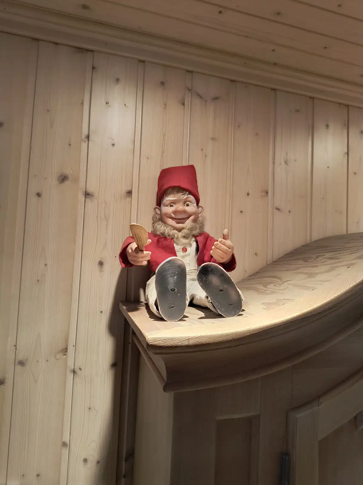 A smiling, bearded figurine wearing a red hat and red jacket sits on a wooden ledge against a wooden wall, holding a wooden…