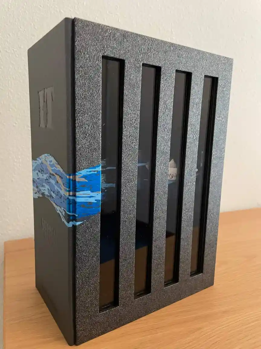 A large, black, textured book with vertical glass panels on its front, resembling a prison cell, sits on a wooden surface…