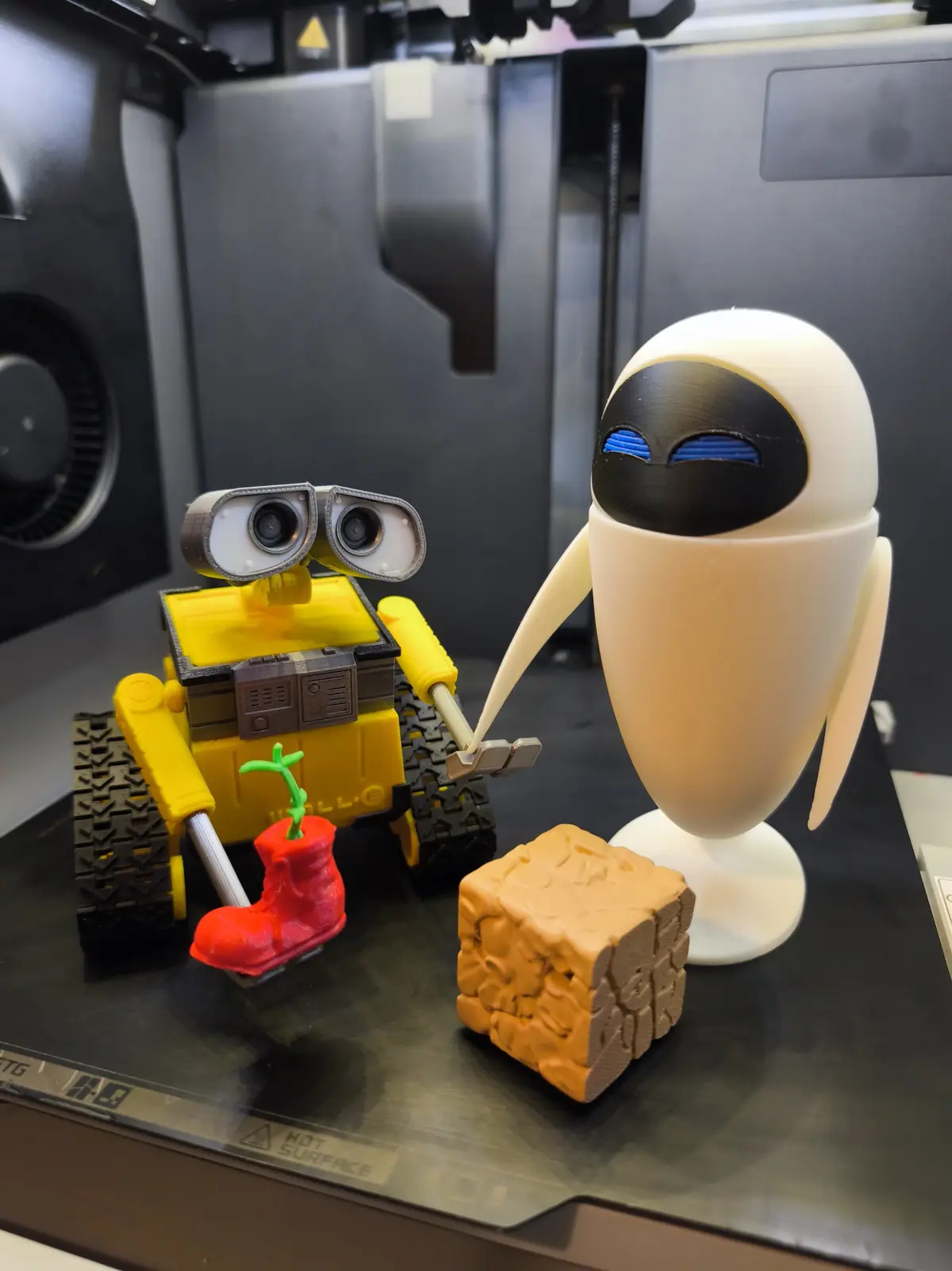 Two 3D-printed robot figurines, Wall-E (yellow, holding a red boot with a green sprout) and EVE (white, with blue eyes)…