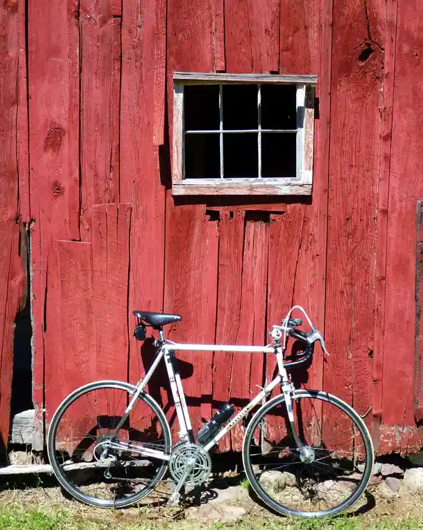 A white road bicycle with black handlebars and seat is leaning against the weathered red wooden wall of a barn, positioned…