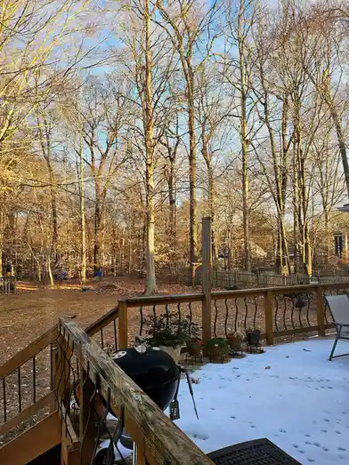 A backyard deck with snow on the ground and a black charcoal grill sitting on the wooden railing. Bare trees with…