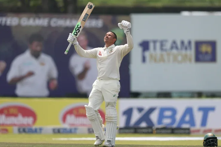 A cricket player in white uniform and protective gear stands on a field, holding his bat high in one hand and raising his…