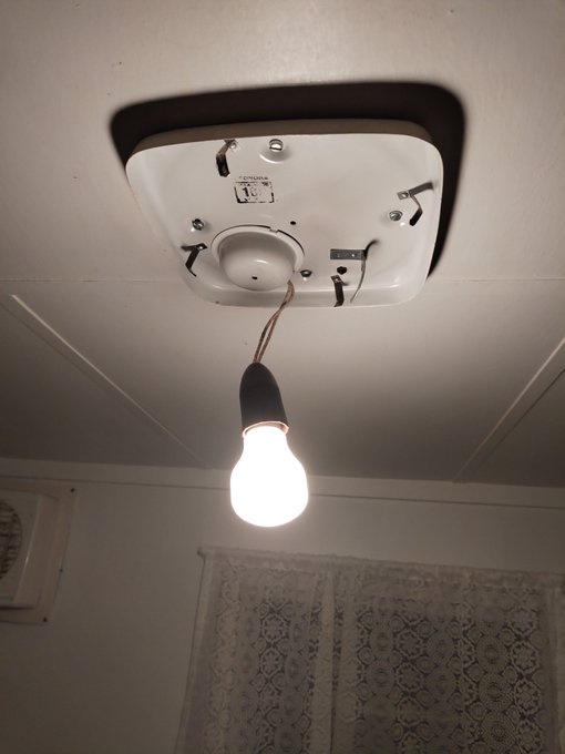 A single lit incandescent light bulb hangs by a frayed wire from a white ceiling fixture with exposed screws and wires, set…