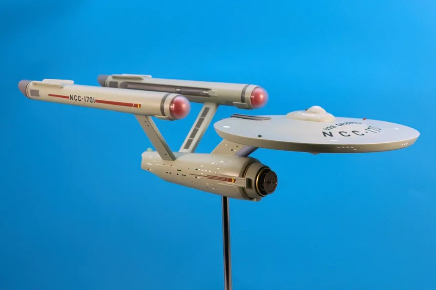 A detailed model of the Starship Enterprise NCC-1701 from Star Trek is displayed on a stand against a solid blue background…