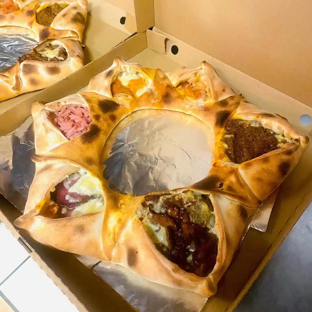 A round, baked flatbread pizza sits in a cardboard box, its center removed and replaced with crumpled aluminum foil, while…