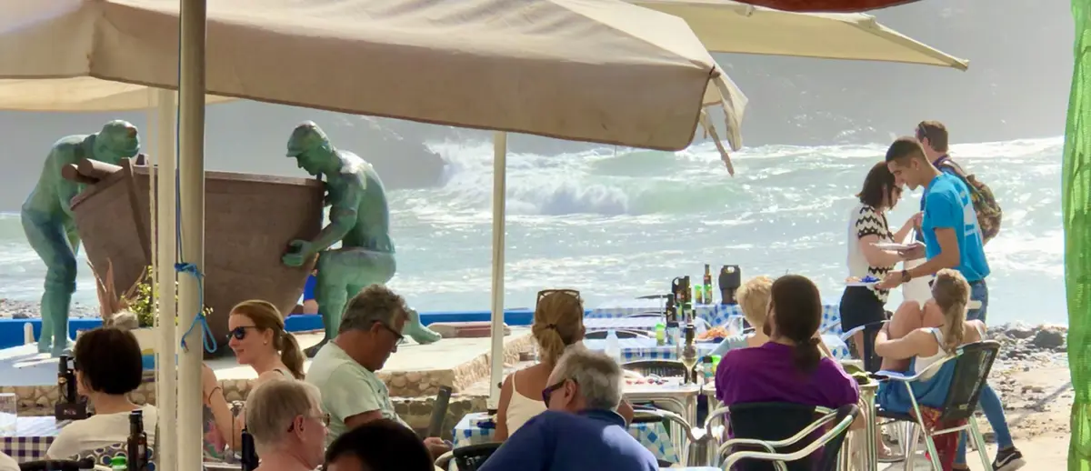 People are seated at outdoor tables under umbrellas at a seaside restaurant, with large green statues of fishermen in the…