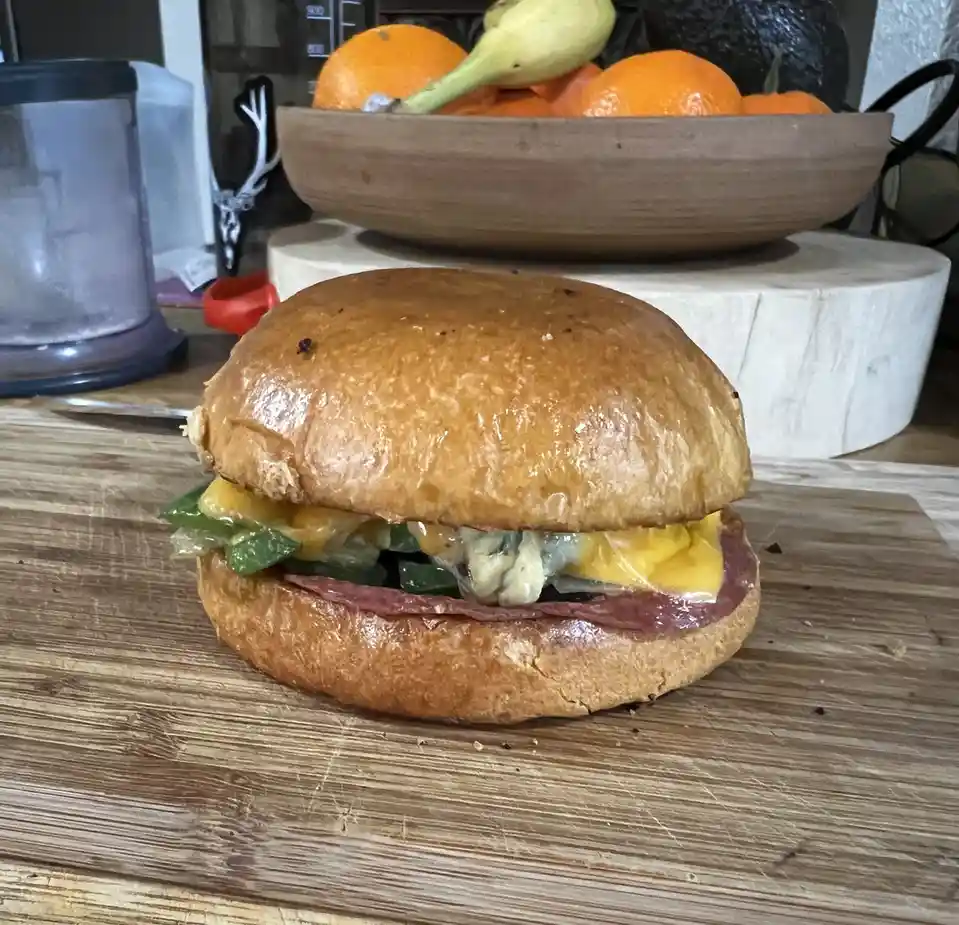 A close-up photo shows a sandwich on a wooden cutting board, with melted cheese, green peppers, and a slice of meat visible…