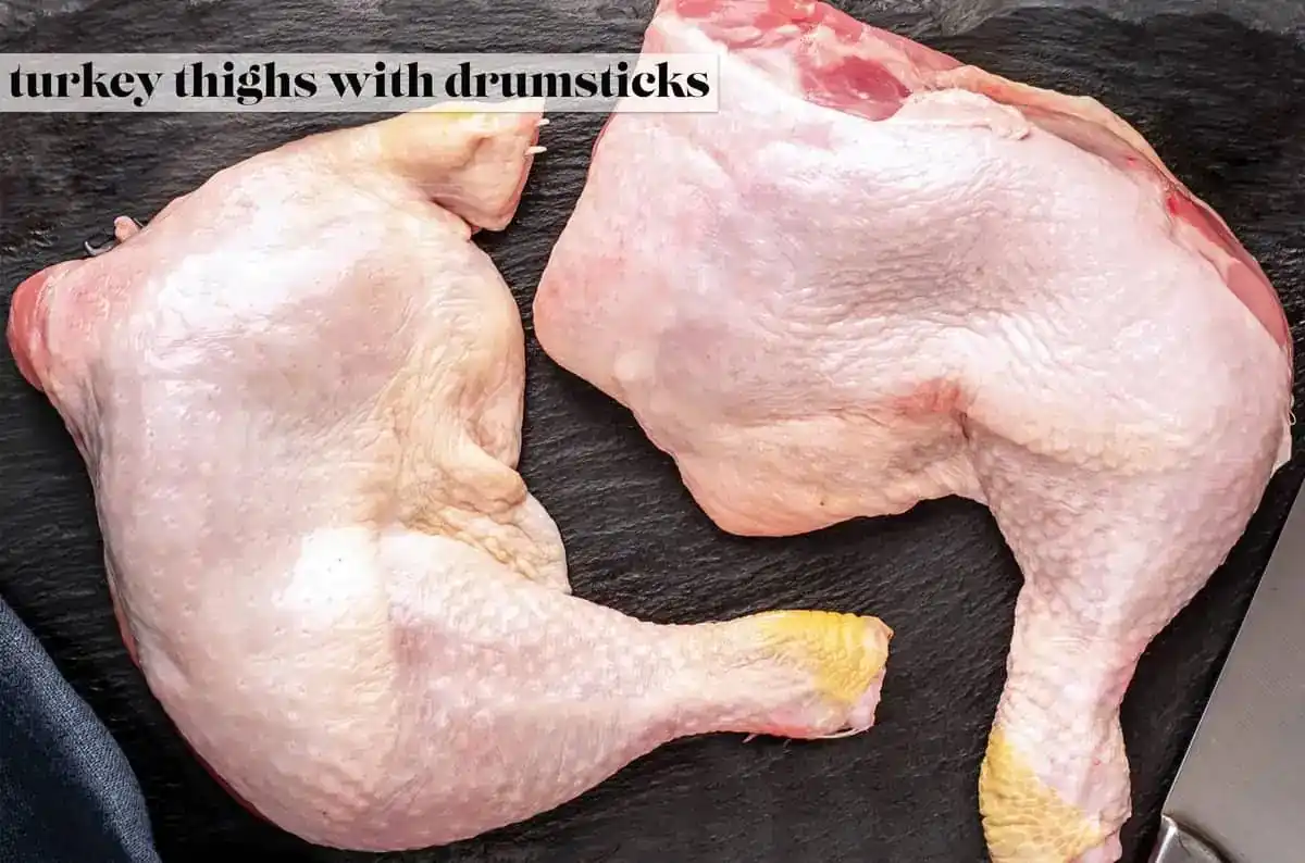Two raw turkey thighs with drumsticks are laid out on a dark slate cutting board, their pale pink skin and yellowish feet…