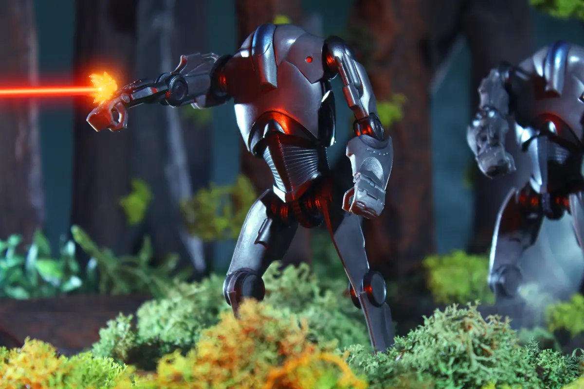 A silver robot action figure is posed firing a red laser from its right hand while standing on mossy ground in a forest…