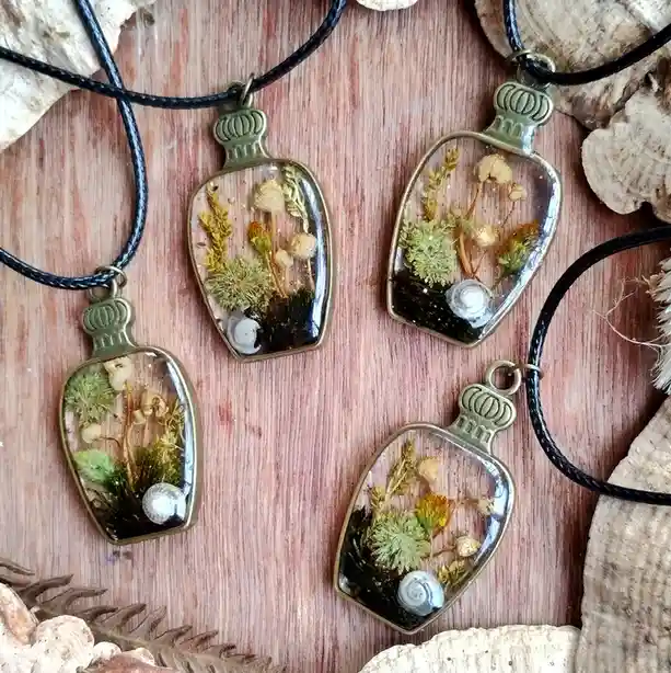 Four pendants shaped like miniature bottles, each containing dried moss, tiny flowers, and a small snail shell, are…