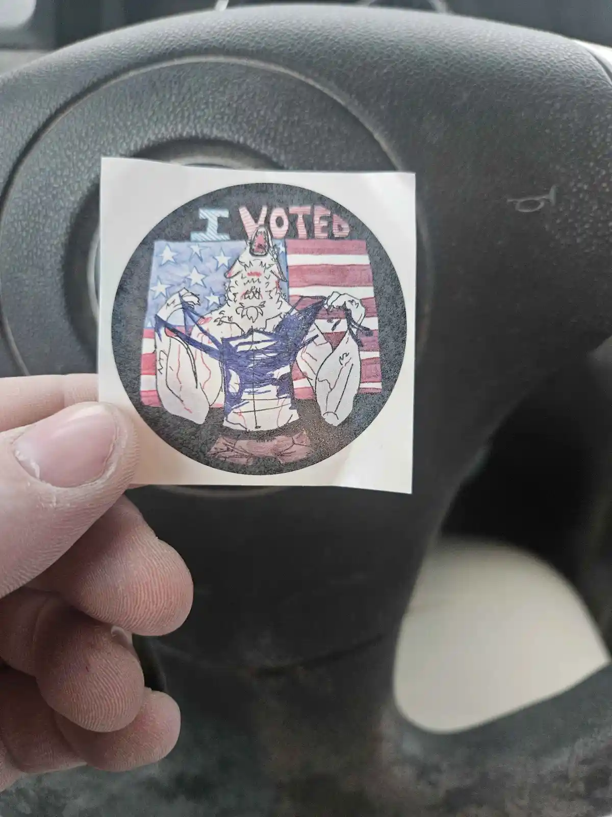 A hand holds a custom-drawn 'I VOTED' sticker in front of a car steering wheel. The sticker features a cartoonish, muscular…