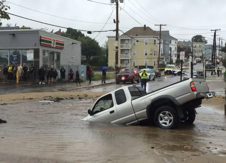 A silver pickup truck is partially submerged in floodwaters on a street, with its front end tilted upward. People are…