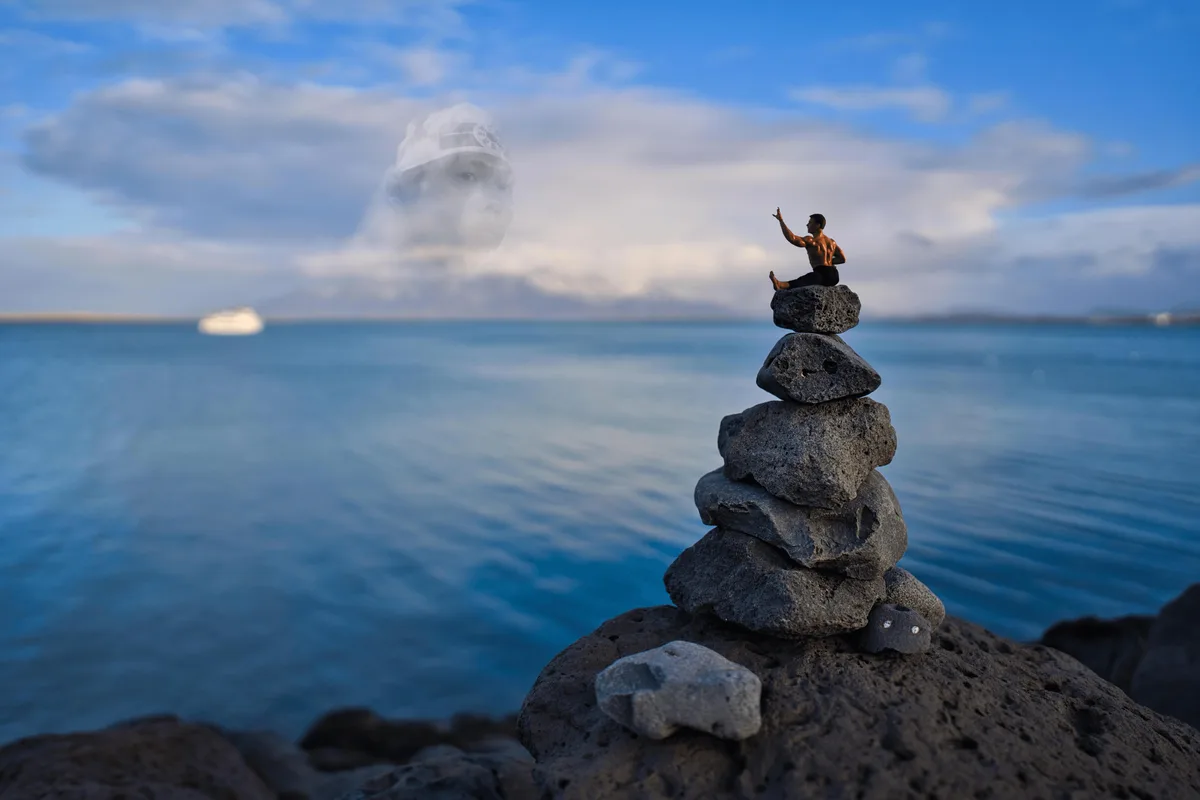 A small figurine of a shirtless man sits atop a stack of dark volcanic rocks on a beach, gesturing upward. Behind him, a…