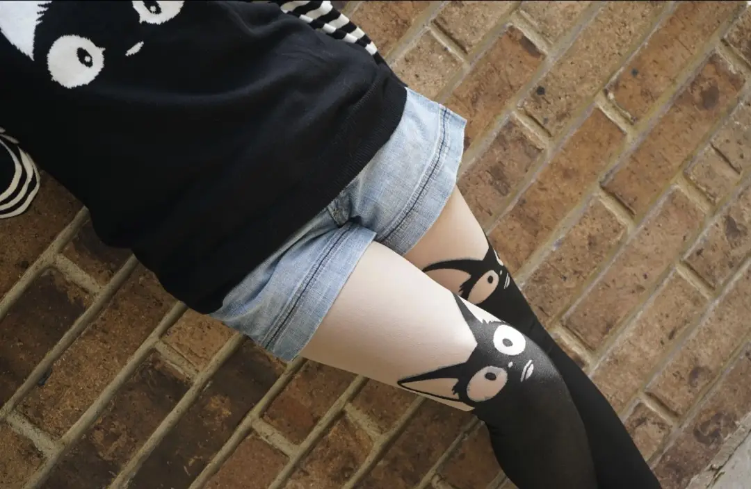 A person wearing a black sweater with a cat face on it, denim shorts, and black tights with cat faces printed on them…