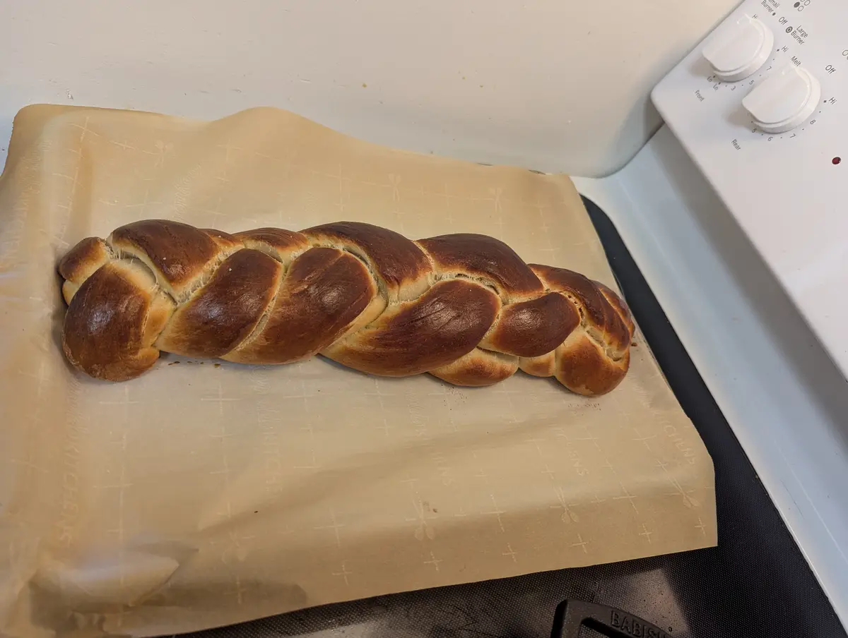 A freshly baked, golden-brown braided bread rests on parchment paper atop a stovetop. The bread has a glossy, evenly browned…