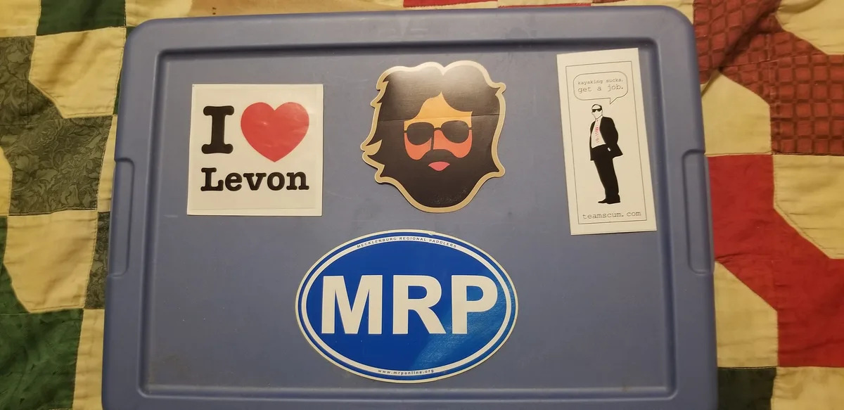 A blue plastic container lid covered in stickers, including 'I ❤️ Levon', a bearded man with sunglasses, 'MRP' (Mecklenburg…