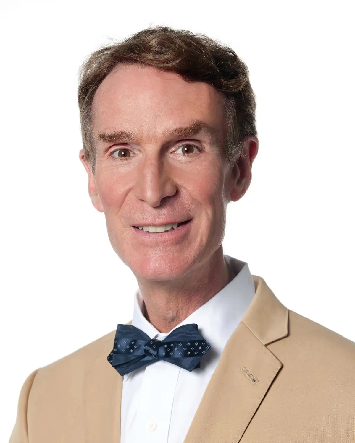 A man with brown hair and a tan suit smiles while wearing a white shirt and a dark blue bow tie with a diamond pattern. The…
