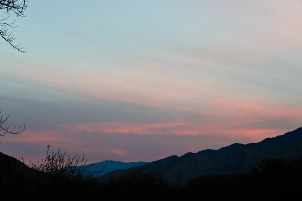 A serene landscape photo of mountains at dusk with pink and blue skies, silhouetted trees and shrubs in the foreground. The…