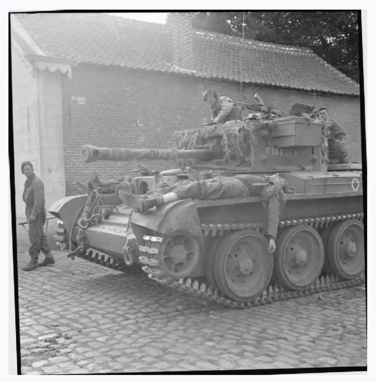 A black-and-white historical photo shows a military tank on a cobblestone street with soldiers. One soldier is lying relaxed…