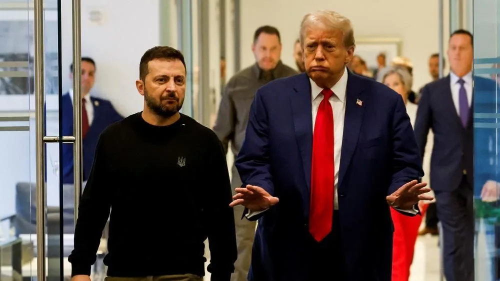 A photo shows two men walking down a hallway; the man on the right, wearing a blue suit and red tie, has his hands out and a…