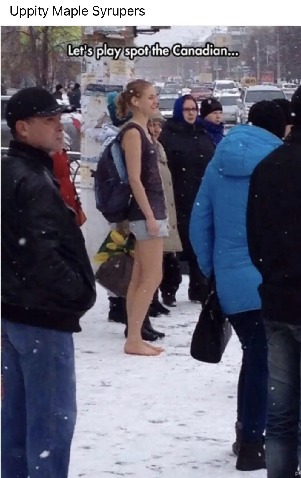 A group of people stand in a snowy urban street, with a young woman in shorts and bare feet standing out prominently. The…