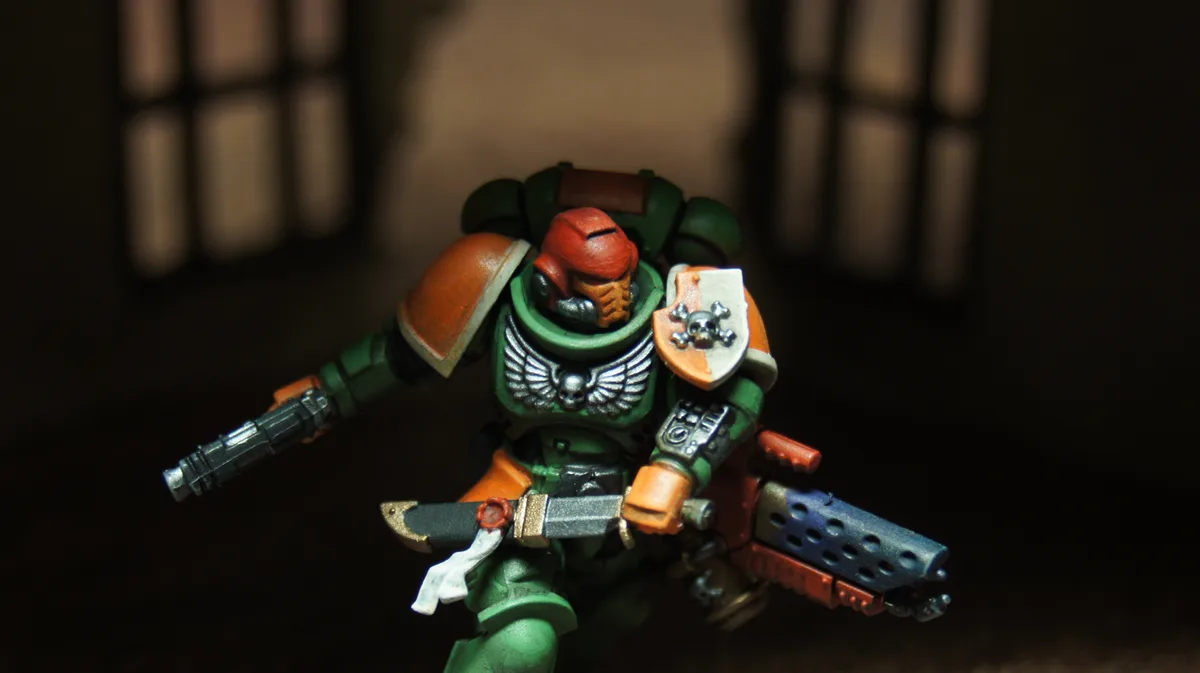 A close-up photo of a painted Warhammer 40,000 Space Marine miniature in green and orange armor, holding a gun and a sword…